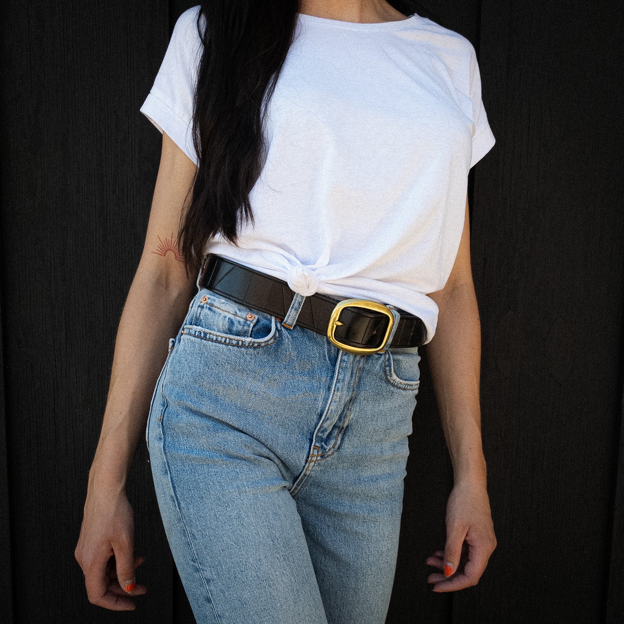 Detail shot of geometric hand-tooling on a black handcrafted leather belt worn by a model with jeans, a sunrise tattoo, and black hair.