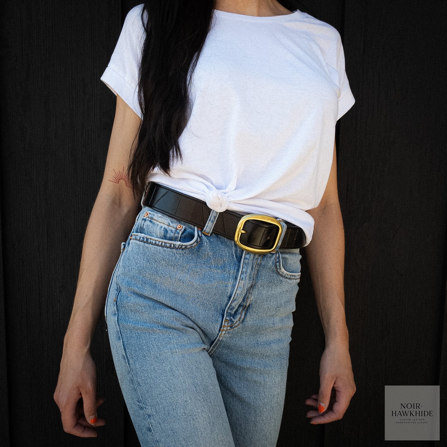 Close-up of the geometric hand-tooled pattern on a black leather belt worn by a model with blue jeans and a white t-shirt featuring a sunrise tattoo.